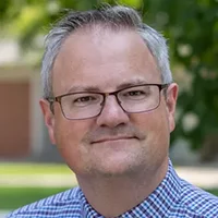 Ryan Harder, a white man wearing glasses and a blue and white checked shirt, smiling against a neutral background